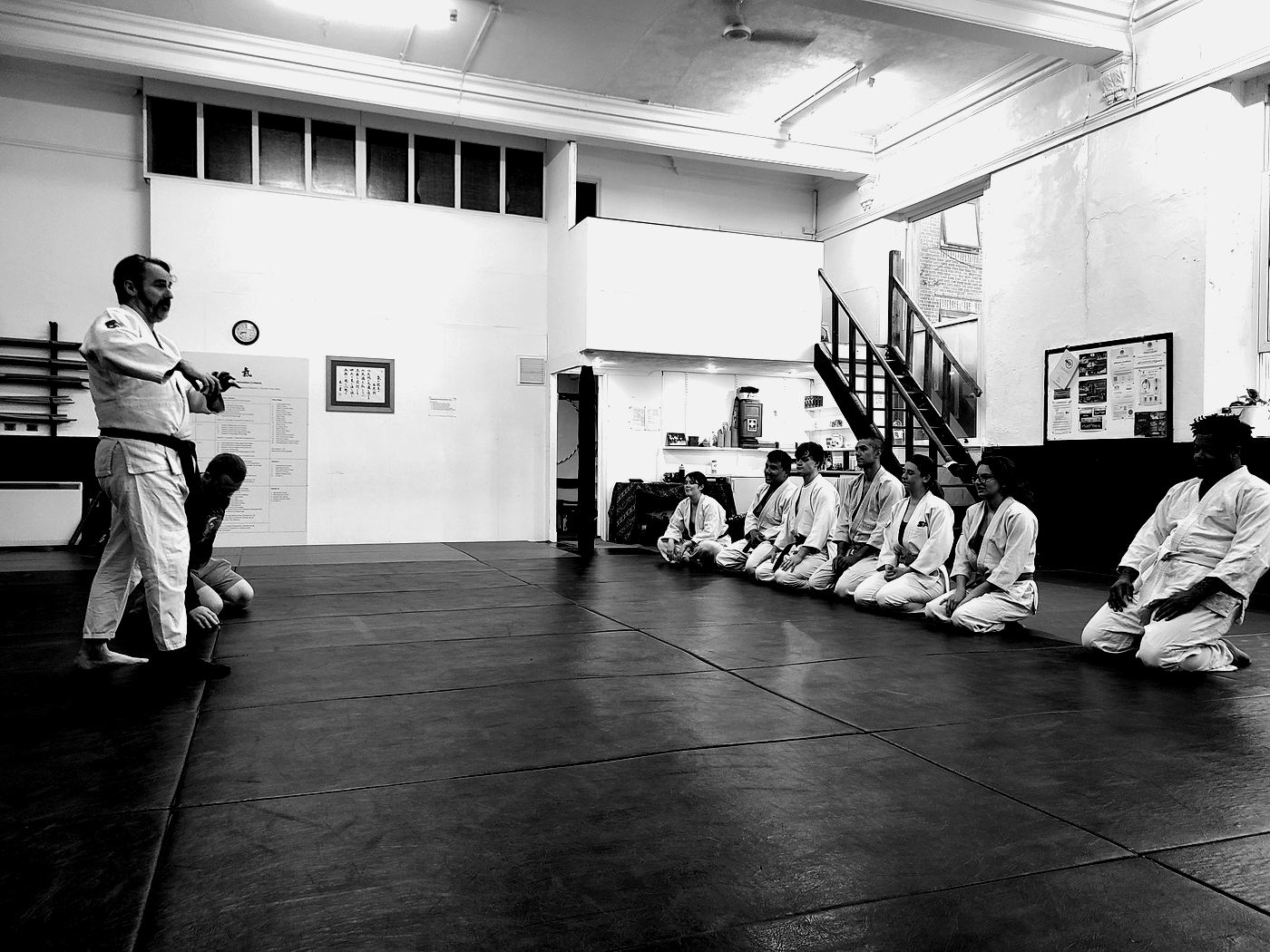 A black belt instructor demonstrates a technique while a row of students kneels on the mat